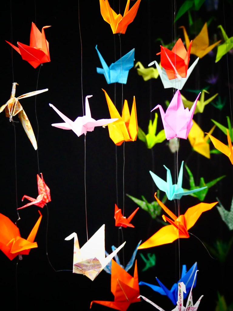 Colourful origami birds hang on strings. Some are white, some are orange, some blue, some green. 