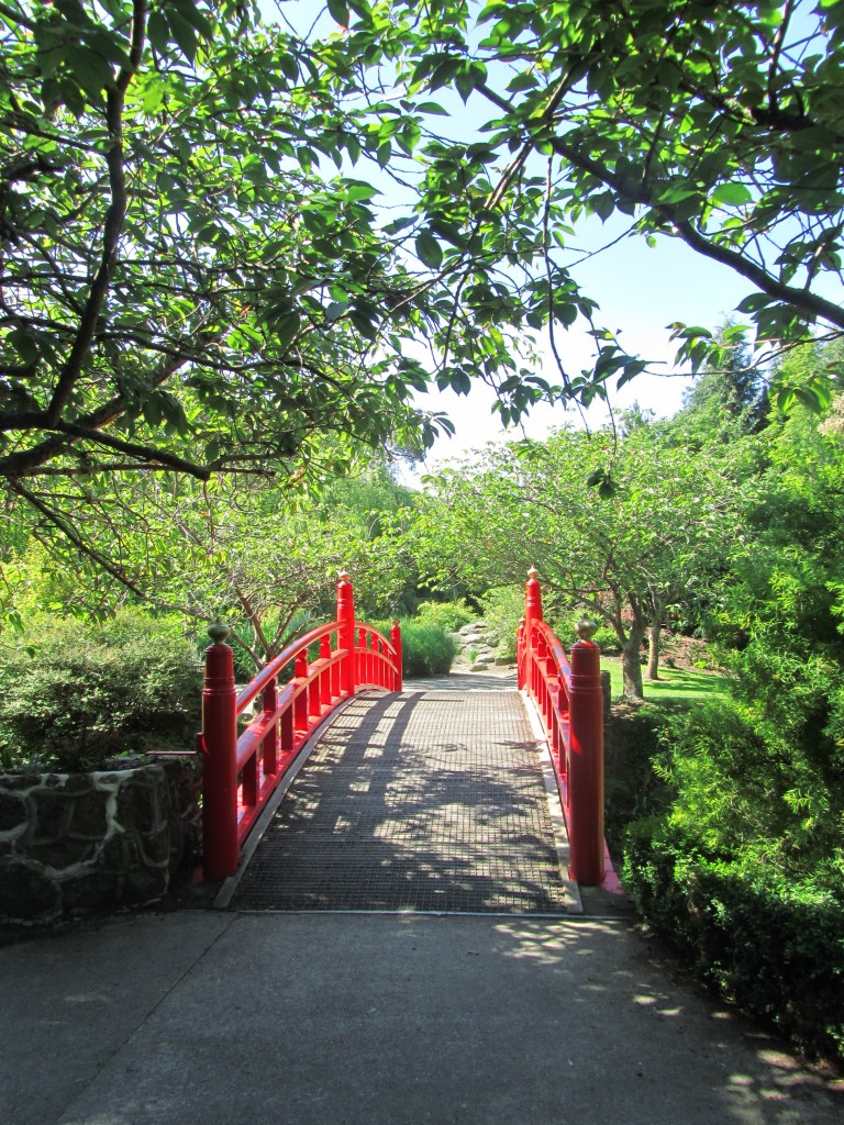 A bridge with bright red handrails curves away into the bright greenery of a botanical garden park. It is bright and sunny, the light of midday.