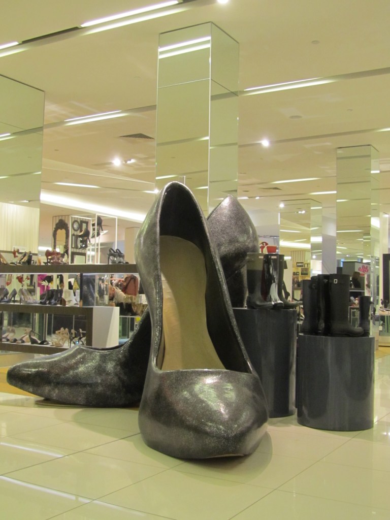 A pair of giant grey high heels in the middle of a large shoe shop's shoe display.